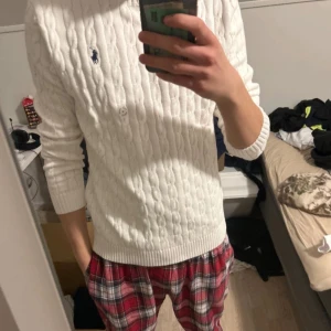 Vit stickad tröja från Ralph Lauren - Säljer en stilren vit stickad tröja från Ralph Lauren med kabelstickat mönster och den klassiska loggan broderad på bröstet. Tröjan har långa ärmar och en rund halsringning. Perfekt för en elegant och avslappnad look.