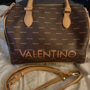 Brun axelväska från Valentino - Snygg brun axelväska från Valentino med logotypmönster. Väskan har en justerbar axelrem och handtag i ljusbrunt syntetmaterial. Perfekt för att bära dina dagliga nödvändigheter med stil.