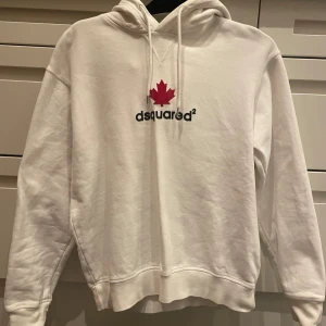 Vit hoodie från Dsquared2 - Säljer en snygg vit hoodie från Dsquared2 med en röd lönnlövsdetalj och logotyp på framsidan. Den har en klassisk passform med långa ärmar och justerbar huva. Perfekt för en stilren och bekväm look. 