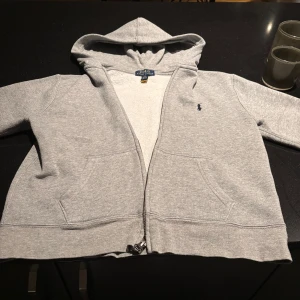 Grå hoodie från Polo Ralph Lauren - Säljer en stilren grå hoodie från Polo Ralph Lauren. Den har en dragkedja framtill och en klassisk huva. Perfekt för en avslappnad look. Den ikoniska loggan är broderad på bröstet. Passar bra till både jeans och joggers. Den är 10-12 år gammal!