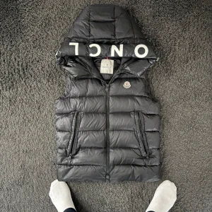 Svart dunväst från Moncler - Snygg svart dunväst från Moncler med huva och dragkedja. Västen har en broderad logotyp på bröstet och text på huvan. Perfekt för kyliga dagar. Den e jättefint skick och har inte använt den alls mycket! Såklart äkta, storlek 1 vilket motsvarar S