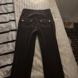 Bruna velourbyxor från Juicy Couture - Snygga bruna velourbyxor från Juicy Couture med broderad logga på fickan. Byxorna har en hög midja med snörning och två fickor fram med knappar. Perfekta för en avslappnad och trendig look. Har andvänt fyra gånger, köpte de för 1200