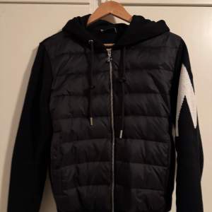 Snygg svart cardigan jacka från Moncler med dragkedja och huva. Jackan har långa ärmar med ett vitt mönster och Moncler-logga på ärmen. Perfekt för kyligare dagar.