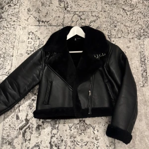 Svart skinnjacka med pälsdetaljer - Snygg svart skinnjacka med mjuka pälsdetaljer och dragkedja. Jackan har en cool design med bälte vid kragen och långa ärmar. Perfekt för att hålla värmen med stil. Jackan är kroppad och är från H&M. Öppen till att mötas upp i Stockholm.