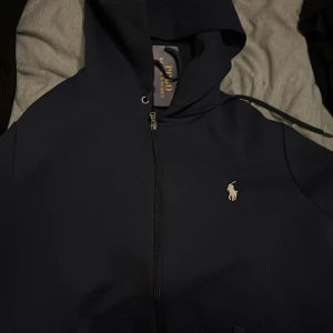 Mörkblå hoodie från Ralph Lauren - Säljer en snygg mörkblå hoodie från Ralph Lauren med dragkedja och den klassiska loggan broderad i vitt på bröstet. Perfekt för en stilren och avslappnad look. 🐎