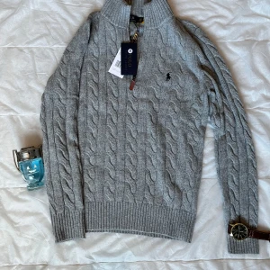  Polo Ralph Lauren quarter zip - Säljer en stilren grå stickad tröja från Polo Ralph Lauren med kabelstickat mönster och dragkedja vid halsen. Tröjan har långa ärmar och ribbade muddar vid ärmslut och nederkant. Perfekt för kyligare dagar!