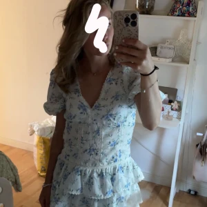 Blommig klänning med volanger - Jättesöt och somrig vit klänning med blå blommor och volangdetaljer. Klänningen har korta puffärmar och en v-ringad hals. Den har använts en gång och är köpt från Vylander, jag säljer den för 400kr. Xs/S🌸🫶🏼 Hör av dig vid fråga!