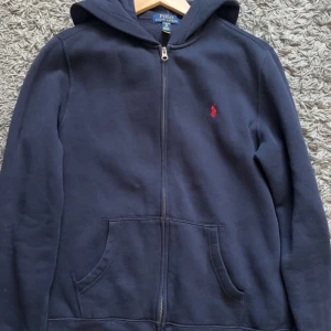 Mörkblå hoodie från Polo Ralph Lauren - Säljer en snygg mörkblå hoodie från Polo Ralph Lauren med dragkedja och huva. Den har en röd broderad logga på bröstet och praktiska fickor framtill. Perfekt för en avslappnad stil. Storlek L men passar M lite stor vid midjan bara 