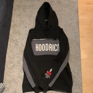 Svart hoodie från Hoodrich - Säljer en svart hoodie från Hoodrich med gråa detaljer och texten 'HOODRICH' tryckt på bröstet. Hoodien har en justerbar huva och en stor ficka framtill. Perfekt för en avslappnad stil.