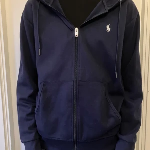 Ralph lauren zip hoodie mörkblå - Mörkblå ziphoodie från Ralph Lauren. Tröjan är i jätte bra skick förutom en extremt liten defekt på vänster arm (se bild 4) man tänker dock absolut inte på det när man har på dig tröjan. Storlek M.