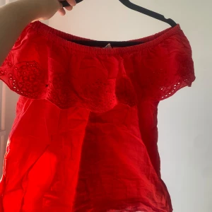 Röd offshoulder topp - Säljer en superfin röd offshoulder topp med vackra spetsdetaljer. Perfekt för sommardagar eller en festlig kväll. Toppen är ärmlös och har en luftig passform som gör den bekväm att bära.