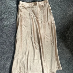Beige långkjol i satin - Säljer en superfin beige långkjol i satin. Den har en elegant och flytande passform som passar perfekt för både vardag och fest. Kjolen är i nyskick och känns riktigt lyxig. Perfekt för höst och vår!