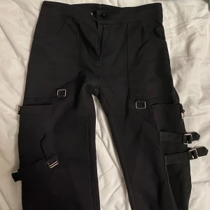 Svarta cargopants med spännen - Ett par supersnygga svarta alternativa byxor som är köpta här på plick! De har en massa detaljer; spännen och fickor. Ser lite ut som trippnyc byxor och är knappt användna. 