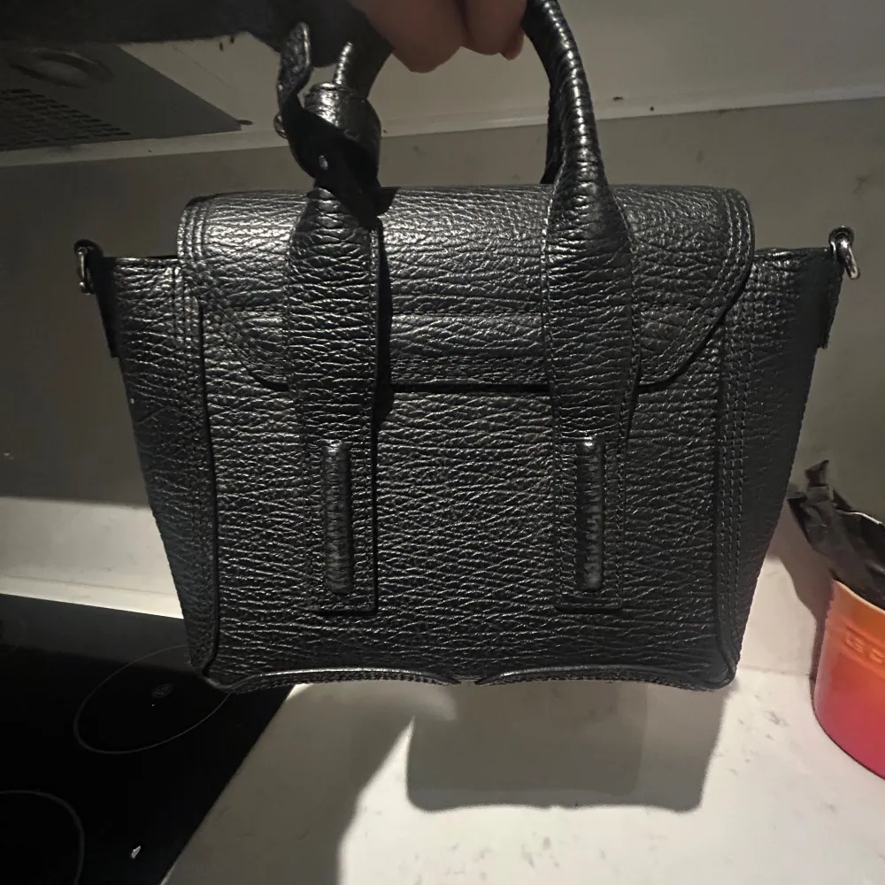 Säljer en snygg svart skinnväska med coola detaljer. Väskan har en praktisk dragkedja och ett stilrent metallspänne framtill. Perfekt för både vardag och fest! Från Phillip Lim. Väskor.