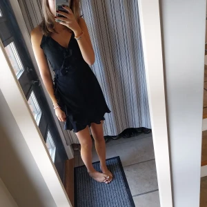 Svart kortklänning med volang - Säljer en superfin svart kortklänning med volangdetaljer. Den har en omlott-design och knytning i midjan som ger en smickrande passform. Perfekt för sommarens fester eller en utekväll! ❤️