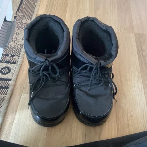 Svarta moon boots - Säljer ett par svarta moon boots i syntetmaterial. De är perfekta för vintern med sin varma och vadderade insida. Skorna har snörning framtill och en glansig yta som ger dem en cool look. Perfekta för kalla dagar!