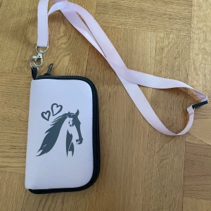 Liten rosa mobilväska  - En rosa mobilväska med häst på framsidan. Den är aldrig använd och i mycket fint skick. Väskan passar en mobil som är 15cm hög och 9,5cm bred.