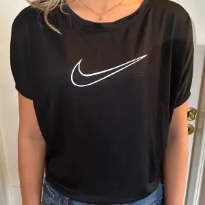 Säljer en svart t-shirt från Nike med den klassiska vita swoosh-loggan på framsidan. T-shirten har en avslappnad passform och korta ärmar, perfekt för både träning och vardag. Materialet känns lätt och andas bra. Passar perfekt till jeans eller träningsbyxor!