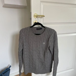 Grå stickad tröja från Ralph Lauren - Säljer en snygg och klassisk grå stickad tröja från Ralph Lauren. Den har ett diskret kabelstickat mönster och en liten broderad logga på bröstet. Perfekt för höst och vinter, med långa ärmar och en bekväm passform. Passar både till vardags och lite finare tillfällen.