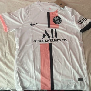 Vit och rosa PSG fotbollströja - Säljer en snygg vit PSG fotbollströja med rosa och svarta detaljer. Tröjan har korta ärmar och är i nyskick. Perfekt för alla Paris Saint-Germain fans! 🏟️