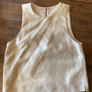 Vit ärmlös topp - Säljer en stilren vit ärmlös topp i ett glansigt material. Perfekt för sommardagar, på kontor eller som en bas i garderoben. Toppen har en enkel design med rund hals och en diskret knäppning i ryggen.