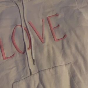 Vit hoodie med rosa text från H&M - Säljer en superfin vit hoodie från H&M med texten 'LOVE' i rosa på framsidan. Den har en klassisk känguruficka och justerbar huva med dragsko. Perfekt för en mysig dag hemma eller en avslappnad dag ute. Passar perfekt till jeans eller leggings!