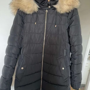 Vinter jacka från Zara - Säljer en svart pufferjacka från Zara i storlek S. Jackan har en mysig fuskpäls på huvan och är perfekt för kalla vinterdagar. Den har en dragkedja framtill och praktiska fickor med knappar. Fodret är gjort av 100% återvunnen polyester, vilket gör de