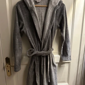Grå långärmad morgonrock - Mysig grå morgonrock med huva och knytskärp i midjan. Perfekt för kalla morgnar eller mysiga kvällar hemma. Den är långärmad och är riktigt bekväm. Materialet är väldigt mjukt och skönt. Älskar den. Knappt använd.