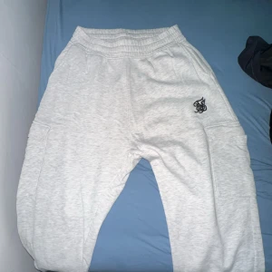 Grå mjukisbyxor från SikSilk - Säljer ett par grå mjukisbyxor från SikSilk i storlek M. Byxorna har en elastisk midja och är superbekväma för chill eller träning. De har en snygg logga på sidan och är i ett mjukt bomullsmaterial. 