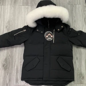 Svart pufferjacka med vit päls - Säljer en supervarm och snygg svart pufferjacka från Moose Knuckles. Den har en stor vit päls runt huvan som verkligen sticker ut. Jackan har långa ärmar och praktiska fickor med knappar. Perfekt för kalla vinterdagar! ❄️