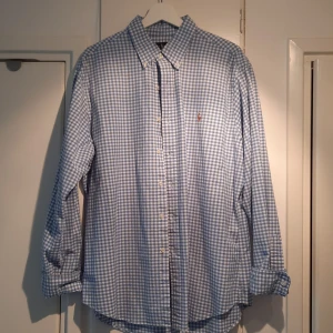 Blå och vit rutig skjorta från Ralph Lauren - Säljer en snygg blå och vit rutig skjorta från Ralph Lauren i storlek XL. Skjortan är slim fit och gjord i stretchigt oxfordmaterial, vilket gör den både bekväm och stilren. Den har långa ärmar och knappar framtill. Perfekt för både vardag och fest! 💙
