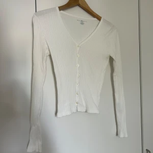 Vit ribbad kofta från Brandy Melville - Säljer en superfin vit ribbad kofta från Brandy Melville. Den har en v-ringad design med knappar hela vägen ner och långa ärmar. Perfekt för både vardag och fest! 🖤Stretchig i material men passar Xs-M
