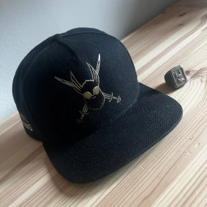 Svart keps med broderad design - Säljer en cool svart keps med en unik broderad design framtill. Perfekt för att ge din outfit en edgy touch. Kepsen har en platt skärm och justerbar passform. Passar både till vardags och speciella tillfällen.