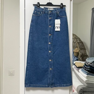 Blå jeanskjol från Zara - Säljer en superfin blå jeanskjol från Zara. Kjolen är midilång och har en rad med knappar framtill som ger en snygg detalj. Perfekt för både vardag och fest! 🦋Köpt för 399kr säljer för endast 179kr.