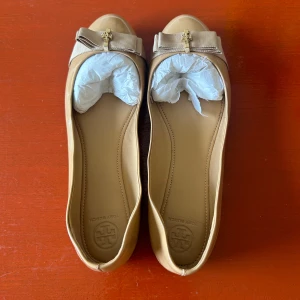 Beige ballerinaskor från Tory Burch - Säljer ett par snygga och eleganta beige ballerinaskor från Tory Burch. Skorna är i skinn och har en fin rosett med gulddetaljer på framsidan. Perfekta för både vardag och fest! Säljer för att jag aldrig använder dem. Som bilderna visar finns några slitningar på dem. Kartongen medföljer.