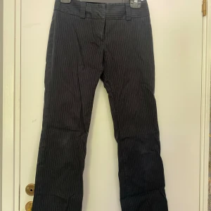 Svarta kostymbyxor med vita ränder - Säljer ett par snygga svarta lågmidjade kostymbyxor med vita ränder. De är gjorda av ett bekvämt bomullsmaterial och har både knappar och dragkedja framtill. Perfekta för både jobb och fest!