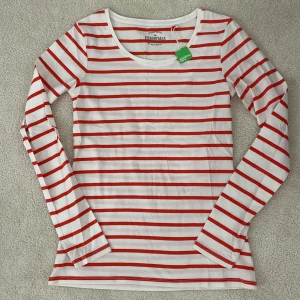 Randig långärmad tröja från The Essentials - Säljer en superfin randig tröja från The Essentials. Den är vit med röda ränder och har långa ärmar. Perfekt för en avslappnad stil och gjord av ekologisk bomull. Helt ny med etikettet kvar!