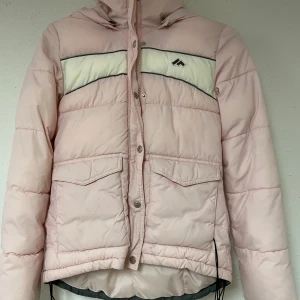 Rosa pufferjacka från Marwin Sports - Säljer en superfin rosa pufferjacka från Marwin Sports. Den har en mysig huva och stängs med både dragkedja och knappar. Jackan har två praktiska fickor framtill och en snygg vit detalj över bröstet. Perfekt för kyliga dagar nu till hösten! 💖