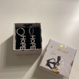 Silvriga örhängen med texten HOPE från IOAKU - Snygga silvriga örhängen från Ioaku med texten HOPE i glittriga bokstäver. Perfekta för att ge en touch av glamour till din outfit. Kommer i en elegant förpackning.