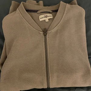 Grå tröja med dragkedja från Hampton Republic - Säljer en stilren beige tröja från Hampton Republic med dragkedja framtill. Perfekt för en avslappnad look. Tröjan har en ribbad krage och är i storlek L.