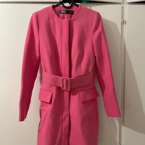 Rosa kappa från Zara - Snygg rosa kappa från Zara med ett matchande bälte i midjan. Kappan har långa ärmar och en stilren design utan krage. Perfekt för att ge en färgklick till din outfit.