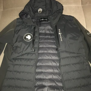 Svart hybrid från Sail Racing limited edition  - Snygg svart pufferjacka från Sail Racing med huva och dragkedja. Jackan har ett broderat märke på bröstet och är perfekt för kyligare väder. Den quiltade designen ger extra värme och stil.