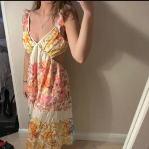 Blommig sommarklänning med öppen rygg - Charmig långklänning med blommigt mönster i rosa, gult och blått. Klänningen har en v-ringad design och en vacker öppen rygg med knytband.
