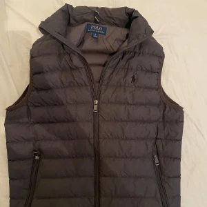 Grå dunväst från Polo Ralph Lauren - Snygg grå dunväst från Polo Ralph Lauren med dragkedja och broderad logga på bröstet. Perfekt för lager-på-lager under kyliga dagar. Västen har en stilren design med praktiska fickor.