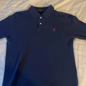 Blå pikétröja från Ralph Lauren - Säljer en klassisk blå pikétröja från Ralph Lauren med röd broderad logga på bröstet. Tröjan har långa armar och är i storlek 14-16 år men sitter som en s väldigt bra pris men kan också diskuteras 