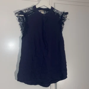 Mörkblå topp från H&M - Fin mörkblå topp från H&M med spetsdetaljer vid axlarna. Toppen är ärmlös och har en rund halsringning med knäppning. 