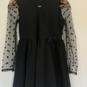 Svart klänning med prickiga ärmar - Fin svart klänning med transparenta prickiga ärmar och omlott-design. Klänningen har en knytning i midjan som ger en fin siluett. Storlek 150 passar 