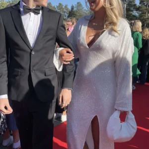 Vit glittrig balklänning - Elegant vit långklänning med glittrande detaljer och djup v-ringning. Klänningen har långa ärmar och en stilren slits framtill, perfekt för tex bal. Endast använd 1 gång. Passar både M och L då den inte är så figursydd. (Jag på bilden är vanligtvis en M)