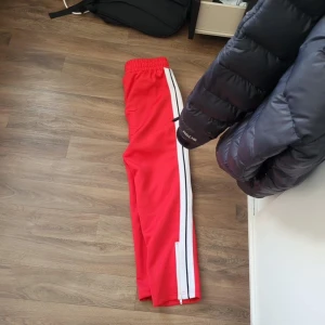 Röda byxor från Palm Angels (Nya) - Snygga röda byxor från Palm Angels med vita och svarta ränder längs sidorna. De har en elastisk midja för extra komfort och en avslappnad passform. Perfekta för en sportig look.