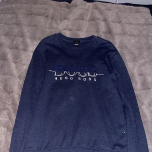 Mörkblå tröja från Hugo Boss - Snygg mörkblå tröja från Hugo Boss med logotypen i vitt och blått på framsidan. Tröjan har långa ärmar och en klassisk rund halsringning. Storlek M, bra skick.